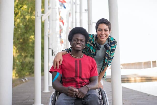 A young man in a wheelchair and a young woman standing next to him, both smiling while on holiday, happy in the knowledge that the man's mobility equipment is protected with wheelchair travel insurance for peace of mind 