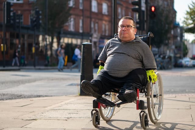 A man in a lightweight folding wheelchair with green gloves pauses on a city street, buildings and a crosswalk in the background.