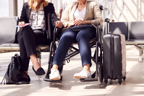 Two women sitting in a waiting area, one in a lightweight folding wheelchair for traveling, with luggage beside them, at an an airport lounge area.