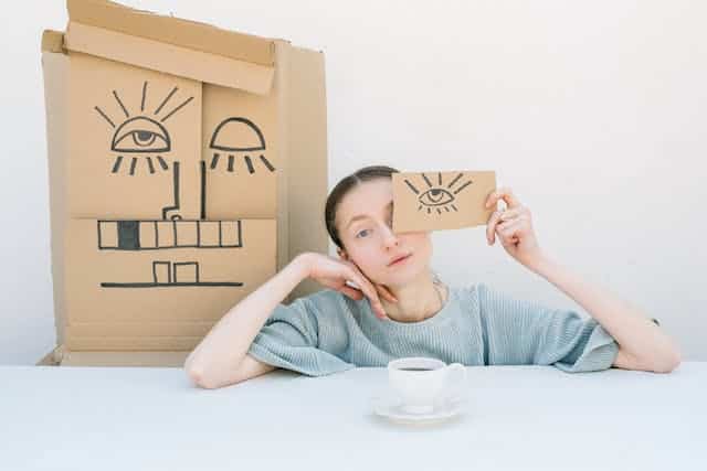A woman with a cup of coffee in hand, holding a cardboard box adorned with a drawing of facial features to represent the experience of thinking or doing things differently to the social norm.