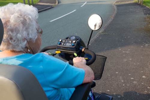An elderly individual drives with insurance for mobility scooters operating a mobility scooter on the roadside with peace of mind knowing she's covered should anything go wrong with her scooter.