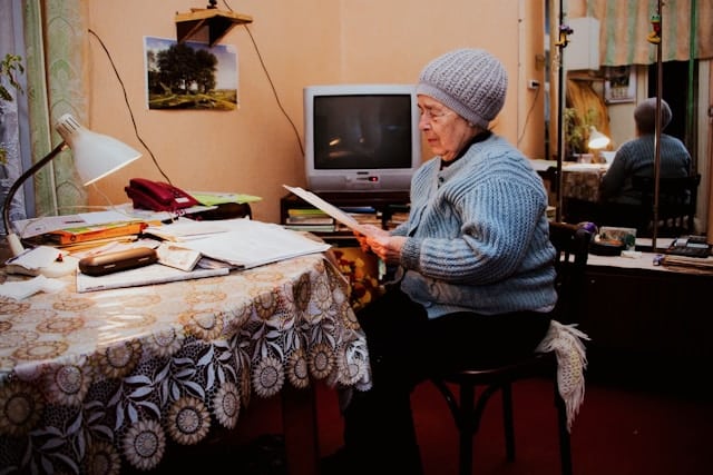 Elderly person reading at a cluttered table, wishing they'd had insurance for their mobility scooter so it could be repaired sooner