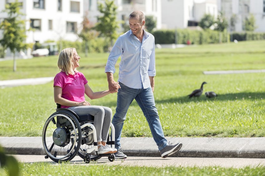 A husband and wife heading out in their neighbourhood using a power assist device on the wife's manual wheelchair for easier maneuverability 
