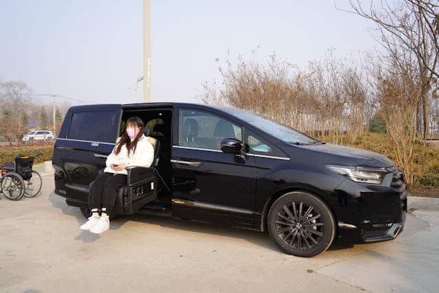 A woman wearing a face mask sits in a mobility seat extending out from a black minivan, with an empty wheelchair nearby, highlighting the accessible transportation options available for people with disability.