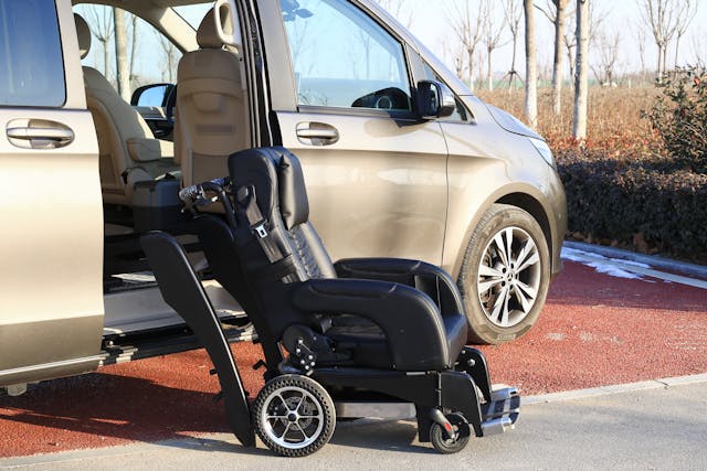 A black wheelchair is positioned next to the open door of a beige van, ready for boarding. In the background, trees and a hedge provide a natural setting for people with disabilities.