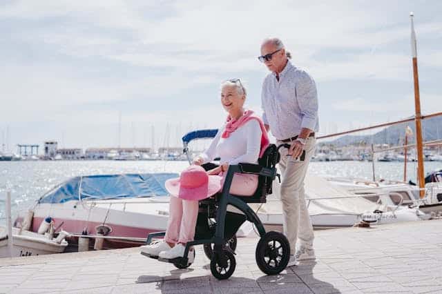 An older adult in a wheelchair, wearing pink, is being pushed by a person standing behind them. They are near a marina with boats docked in the background on a sunny day. Scenes like this remind us of the importance of accessibility and fighting against disability discrimination to ensure everyone enjoys such moments.