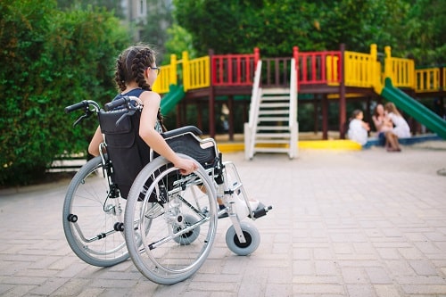 A girl with braids and glasses sits in a paediatric wheelchair in a children's playground.