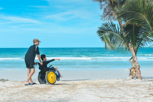this boy is accessing a beach with a wheelchair pushed by his father
