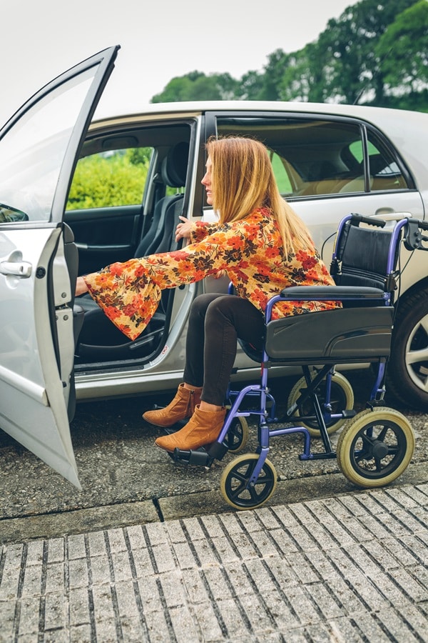 A woman in a floral shirt and boots is seated in a wheelchair, opening a car door as she prepares to transfer into the vehicle, possibly contemplating the best options for comparing disability car insurance.