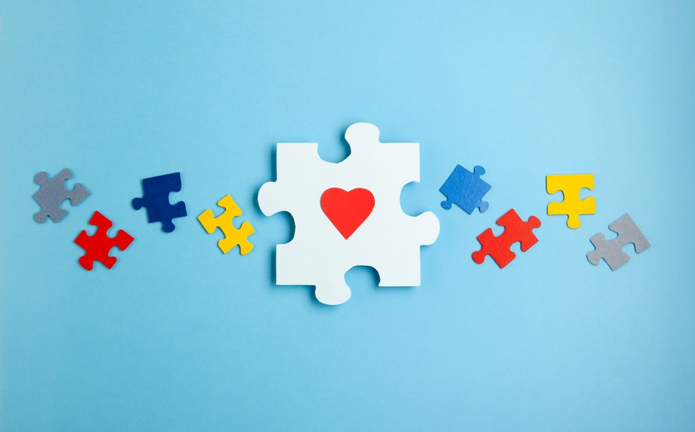 A large white puzzle piece with a red heart in the center is surrounded by smaller colored pieces on a blue background, symbolizing love and unity for International Aspergers Day.