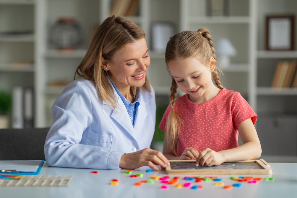 A woman in a white lab coat sits beside a young girl in a pink shirt as they work on a puzzle with colorful pieces, highlighting support for children with Asperger's syndrome on International Aspergers Day.
