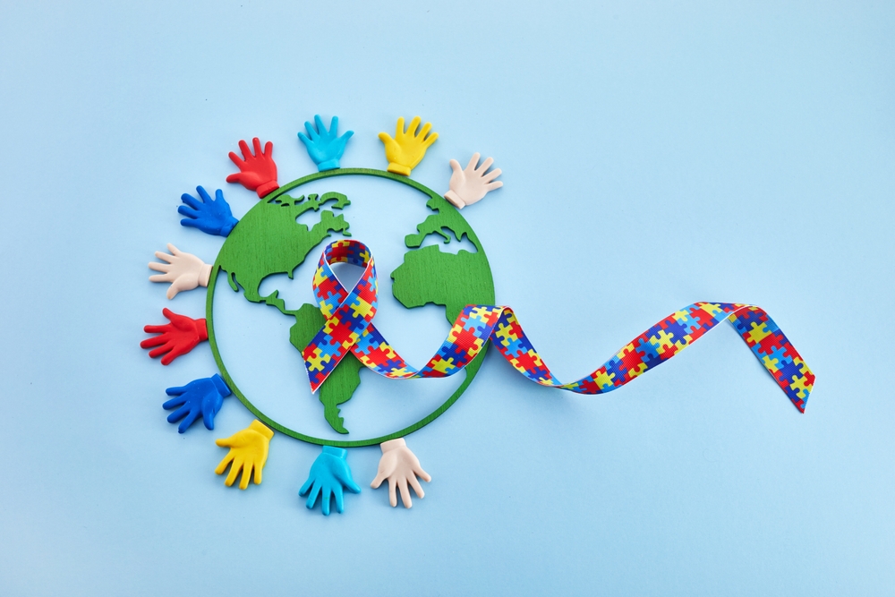 A cutout of Earth surrounded by colorful hand shapes with a puzzle-patterned ribbon, symbolizing autism awareness and Asperger's syndrome, on a light blue background.