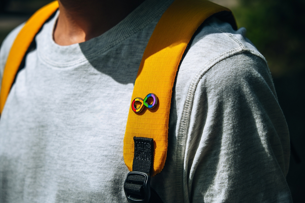 A person wearing a light gray t-shirt and a yellow backpack strap with a rainbow infinity pin attached, showing support for International Aspergers Day and raising awareness for Asperger's syndrome.