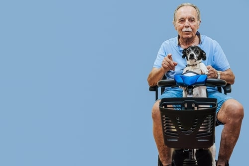 A man in a light blue shirt and jean shorts sits on a mobility scooter, holding a small dog. The background is plain blue. The scene subtly emphasizes the convenience and freedom scooters bring to daily life.