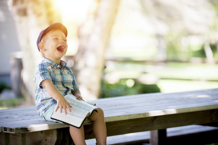 this laughing young boy sitting on a bench reading a book with accessibility symbols is part of the early intervention program