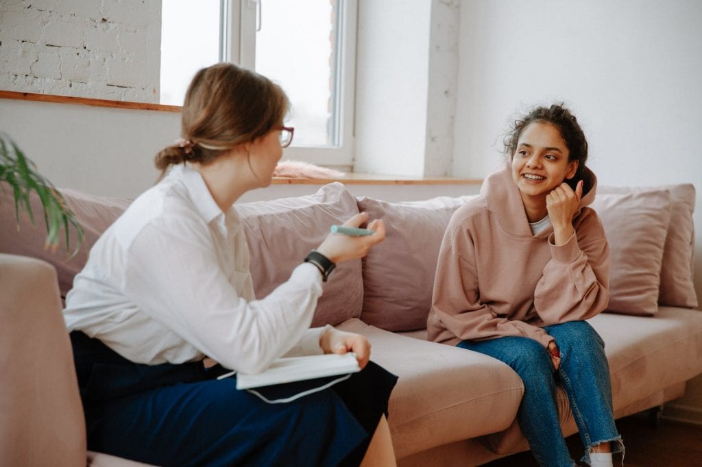 two women meeting for the NDIS - sitting on a couach, one with a notebook and pen and one in a hoodie and jeans