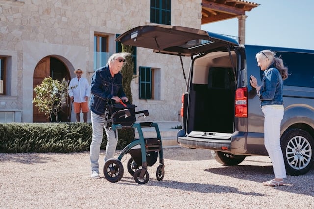 Two elderly people are loading a mobility walker into the back of a van parked in accessible parking bays, while another person stands in the background near a building entrance.