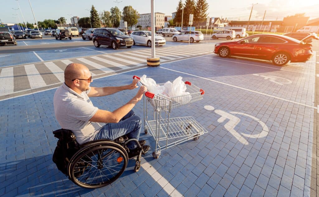 Man in a wheelchair pushes a cart towards a car in a supermarket parking lot.