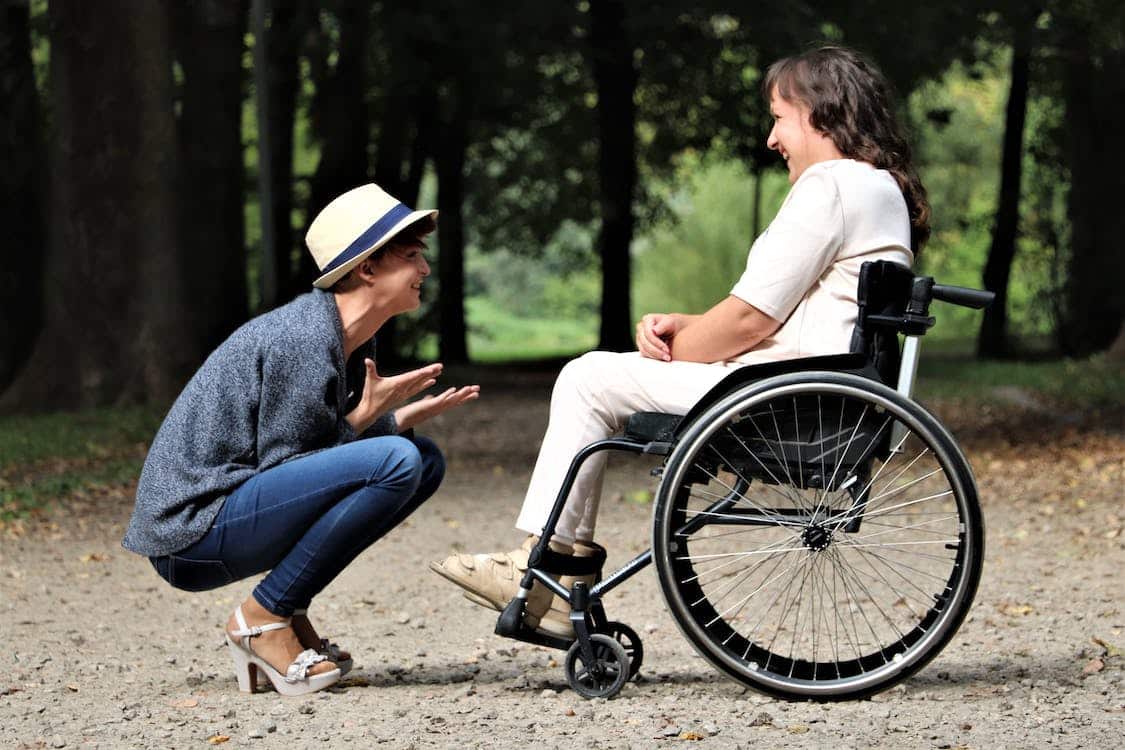 Woman with hat and jeans crouches in front of woman dressed in white in a wheelchair, who's using her disability parking permit in another state