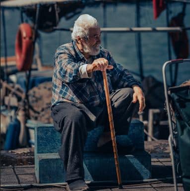 older man in dark flannelette shirt and tracksuit pants sits by the waterside with walking tick in hand after using his disability parking permit insterstate 