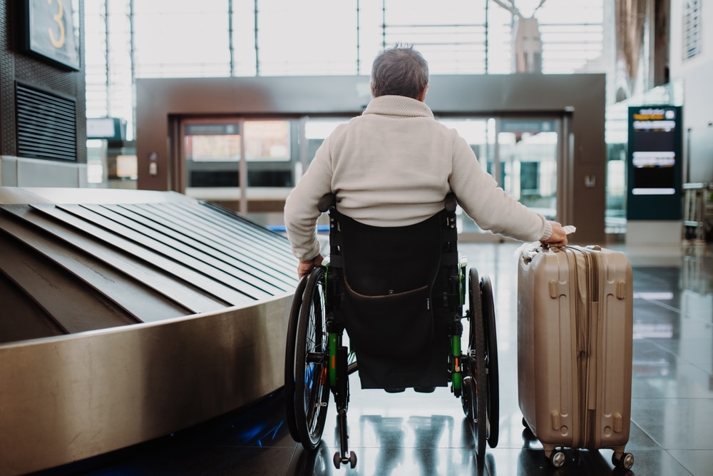 A person in a wheelchair at an airport baggage claim, waiting next to a conveyor belt with a suitcase, feeling relief they have the benefits of wheelchair travel insurance.