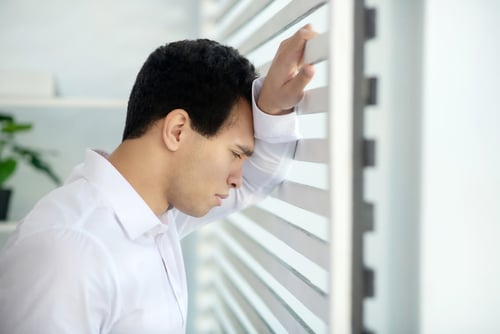 young man with head on arm leaning against blinds in office after stress or anxiety