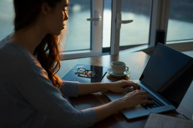 young brunette woman sitting at laptop with coffee cup and glasses to her side