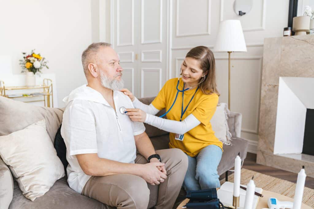 This lady in a yellow shirt is checking a man's heartbeat as they sit on a couch in his lounge room and discuss travelling tips as a carer.