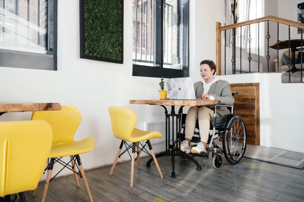 Disability in the workplace is being experienced by this short haired woman in a wheelchair pulled up to a small table with a yellow chair behind it, who is working on her laptop.