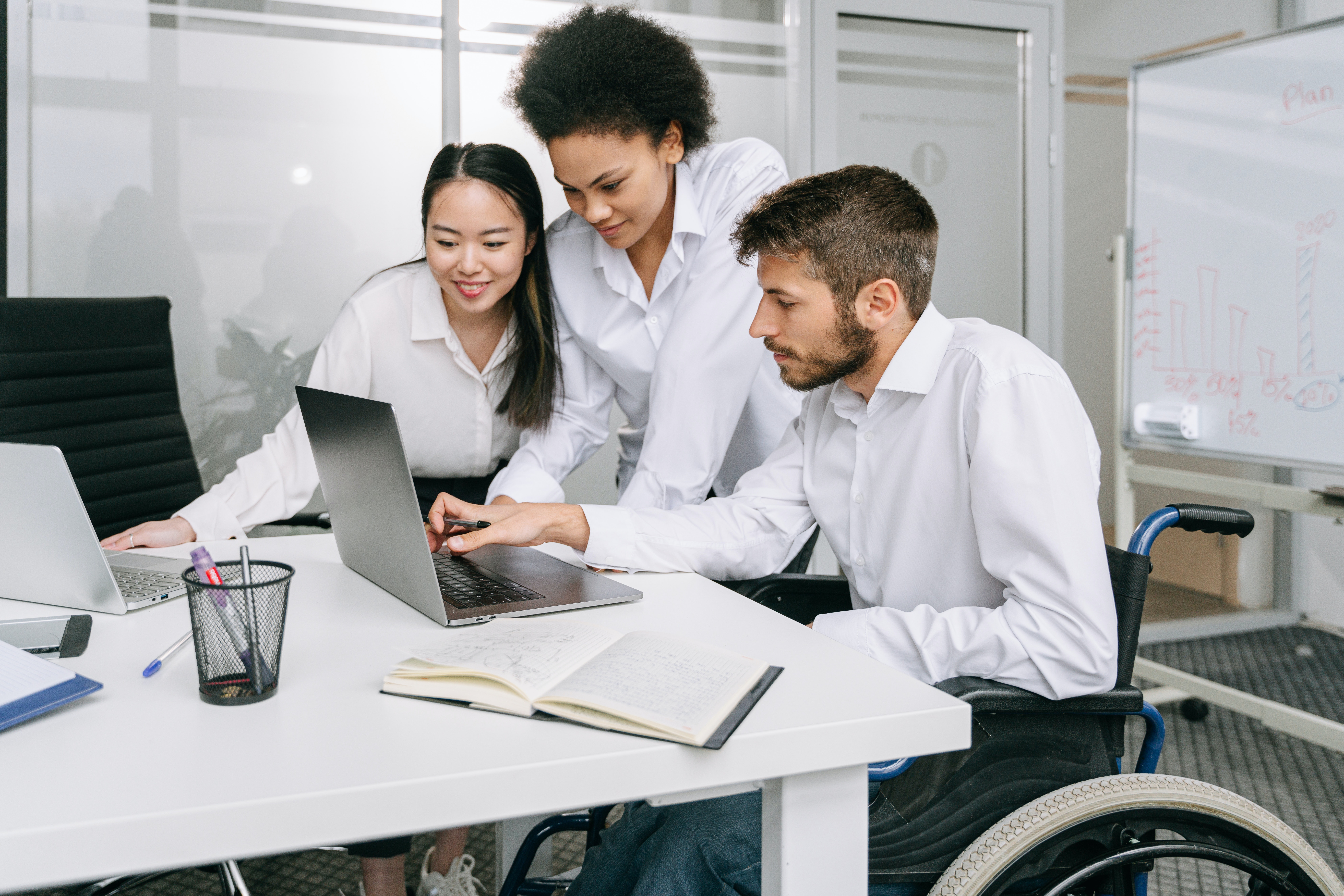 Two female and one male colleague, who is in a wheelchair, look at the same computer screen while discussing disability in the workplace. They all have dark hair and white shirts.