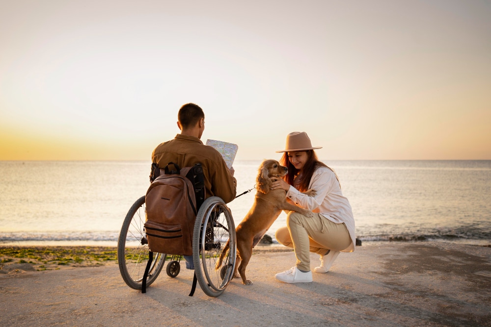 A man in a wheelchair, his carer and assistance dog at the beach together, glad they did their research on travelling with a carer.