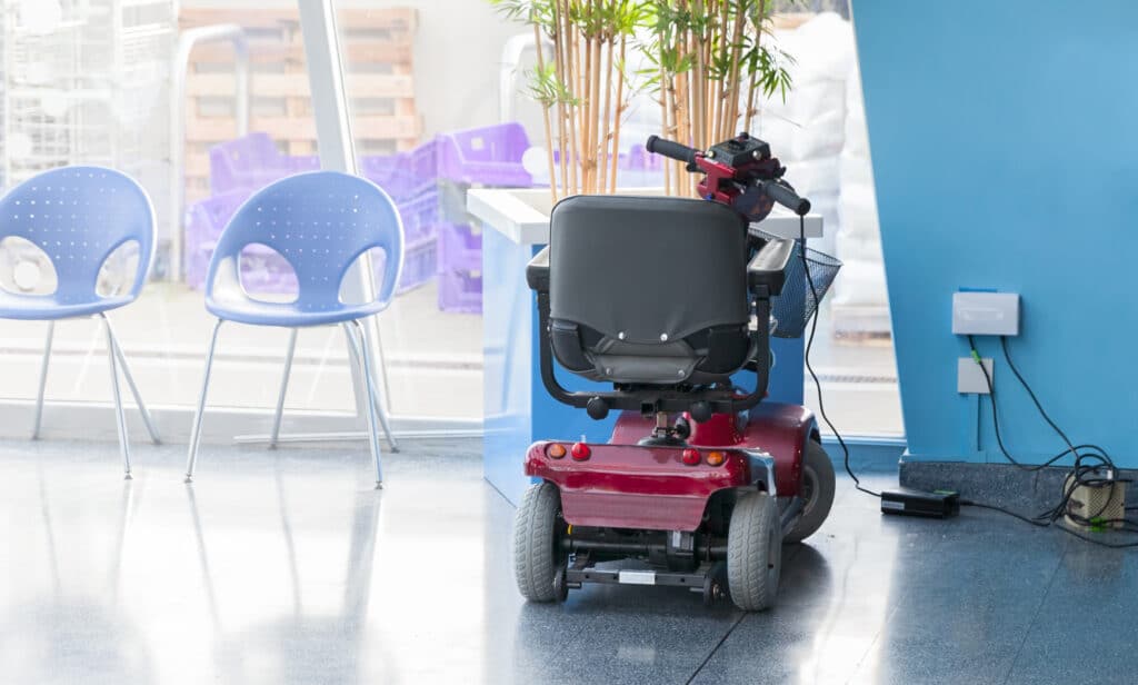 Charge your mobility scooter battery like the one in this black and maroon scooter on charge with chairs and a pot plant around it. 