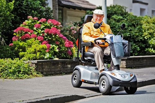 Don't be caught off guard by a dead mobility scooter battery. Be like this man in a mustard jacket with a hat on his scooter in front of pink flowers. 