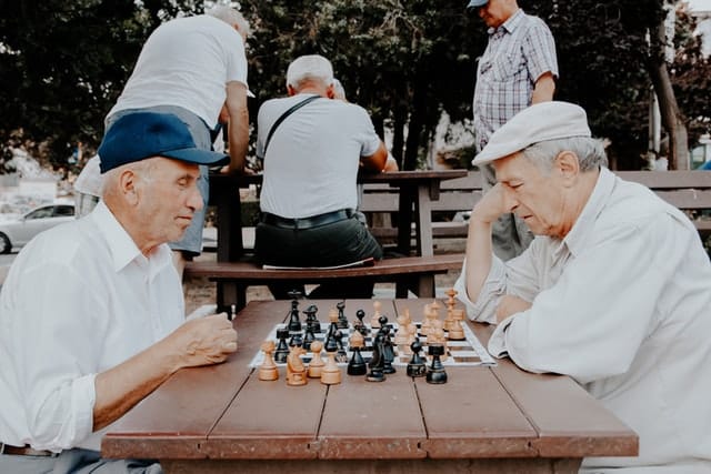 These two older hatted men playing chess at a brown table in a park maintain brain health to help safeguard against dementia