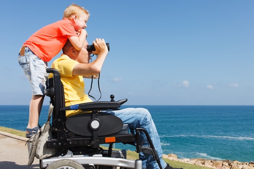 Boy and father in wheelchair look out at the ocean and dream that one day there will be a cure for paralysis