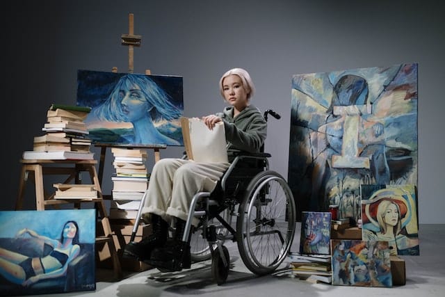 a painter with disability sits in her wheelchair amidst several of her works of art