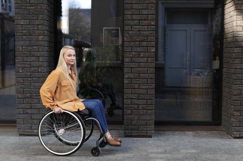 this girl in an orange top and blue jeans in a wheelchair outside a dark brick building deals with chronic pain