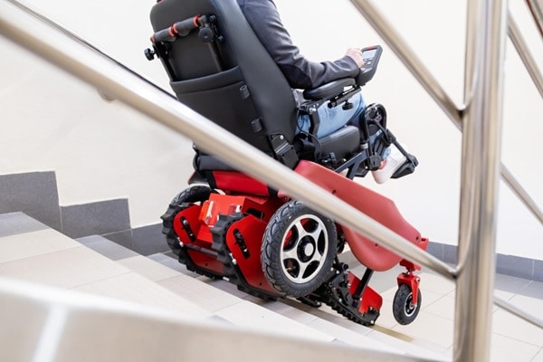 A person in a motorized wheelchair with tracks, similar to a mobile scooter, is ascending a staircase, viewed from behind a stair railing.