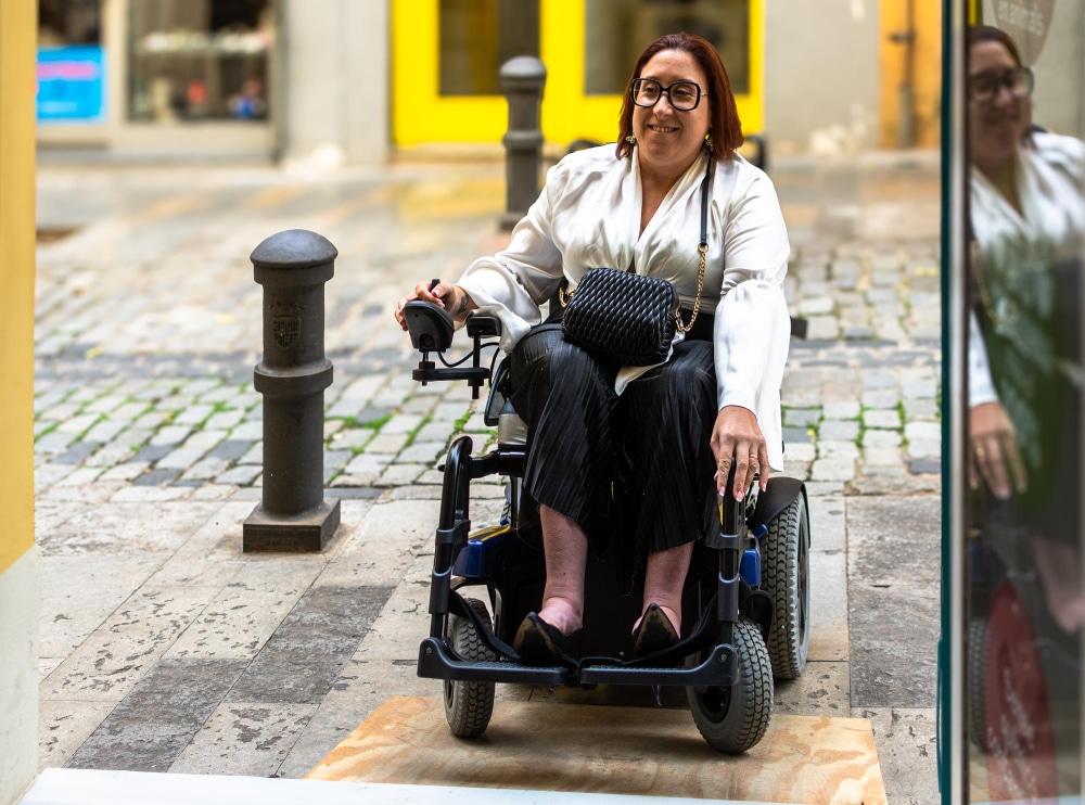 A woman in a motorized wheelchair approaches a building entrance from a cobblestone street, expertly navigating her mobile scooter over a wooden ramp—reminding us of the importance of cleaning mobility scooters for smooth rides.