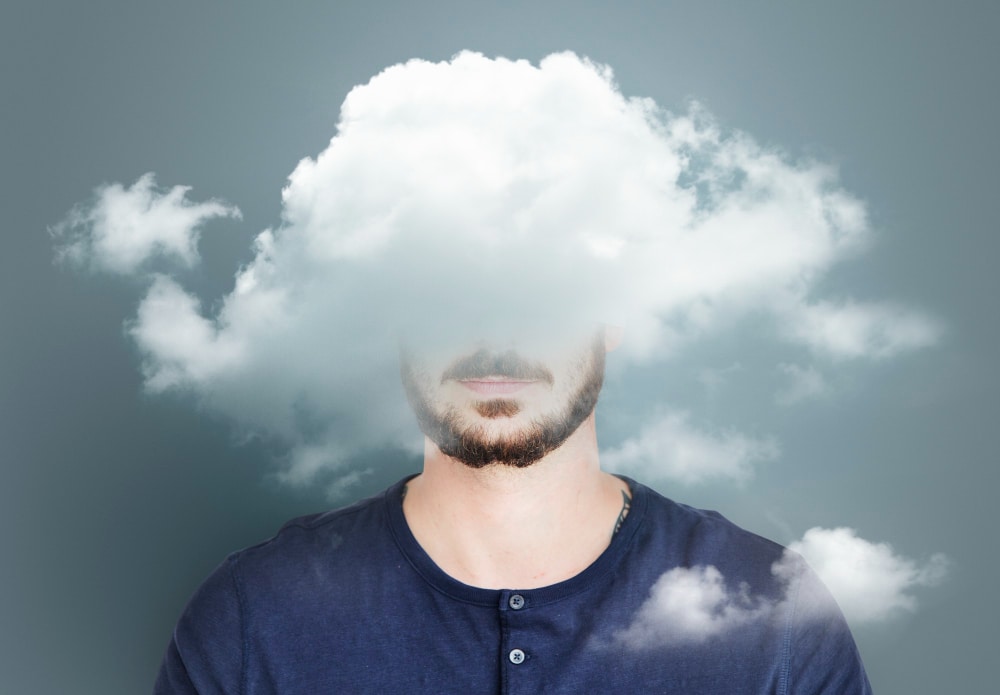 A man with a beard and mustache stands against a gray background, his face digitally obscured by a cloudโevoking the question: what is brain fog, and how can you get rid of it?.