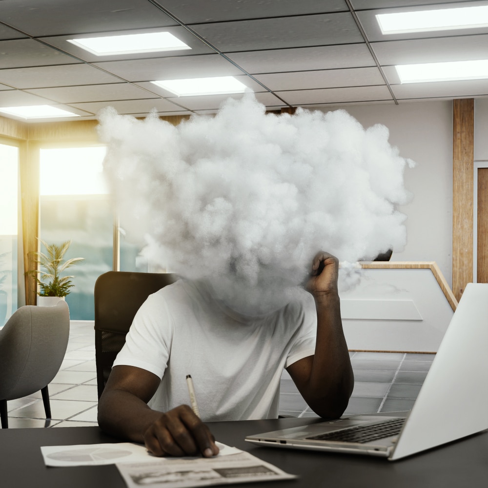 A person sits at a desk with papers and a laptop; their head is replaced by a cloud, visually representing what brain fog feels like in an office setting.