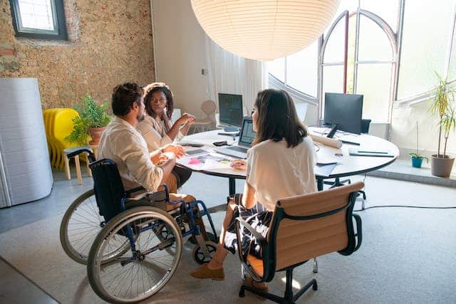 a man using a wheelchair sits at an office table discussing work with two of his colleagues