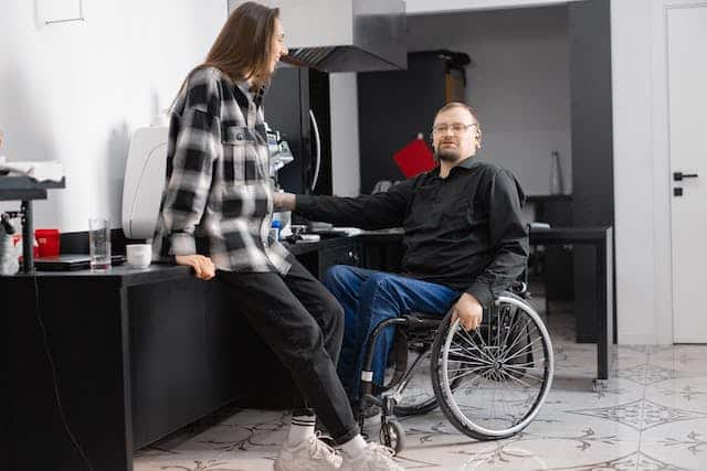 A man in a wheelchair takes a break from filling out an application for an accessible parking permit to have coffee with a colleague in the work kitchenette. 