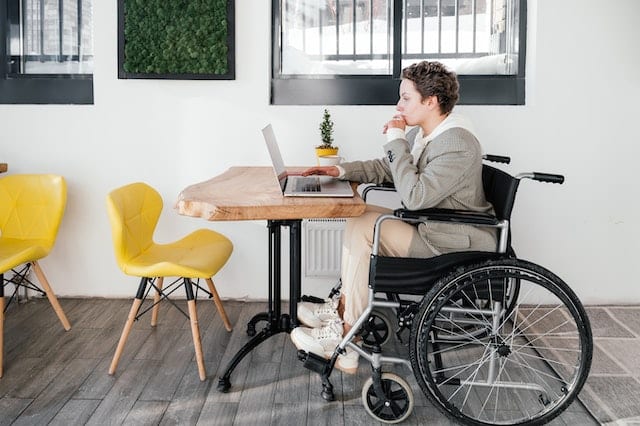 A woman in a wheelchair works on her laptop at a cafe with brightly coloured, yet minimalist furniture. 