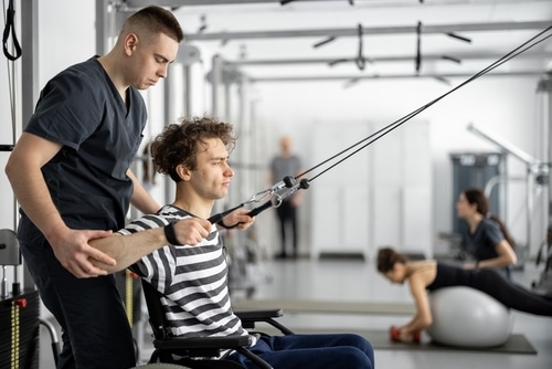 A man in a wheelchair does a wheelchair workout to strengthen his core muscles with the assistance of a physical therapist at a gym.