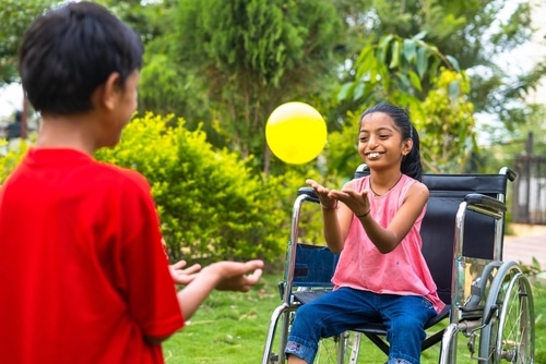 A girl in a wheelchair is playing with a yellow ball, enjoying some wheelchair exercises.