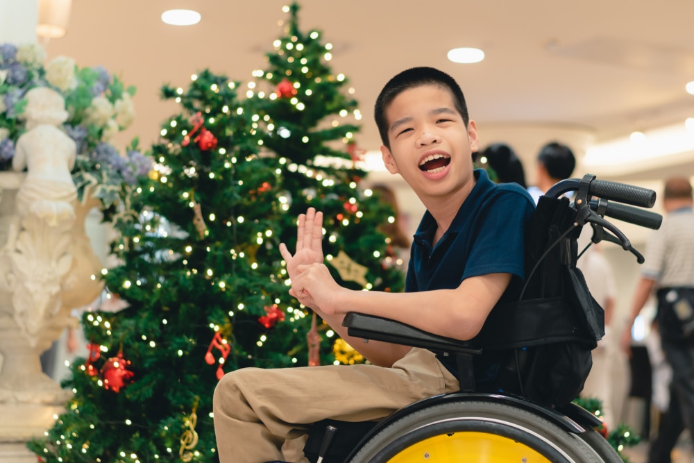 An Australian boy in a wheelchair waves alongside a big Christmas tree filled with gifts for people in wheelchairs