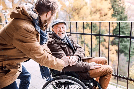 young man in brown jacket looks at and hold the hand of a happy senior man in a brown jacket in a wheelchair