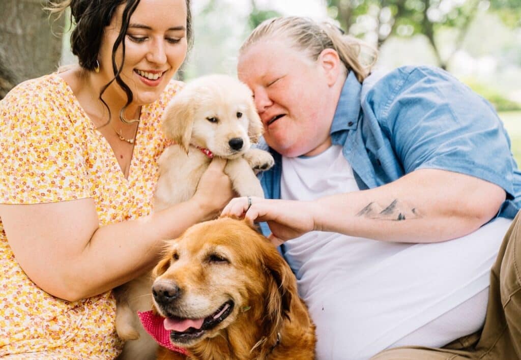 A support worker with a woman who lives with autism sit with their Assistance Dog and their puppy on a blanket in a park. Both the dog and puppy have the protection of service dog pet insurance to help pay for vet costs