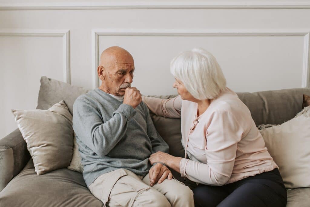 An elderly couple, with limited mobility, sitting on a couch together.